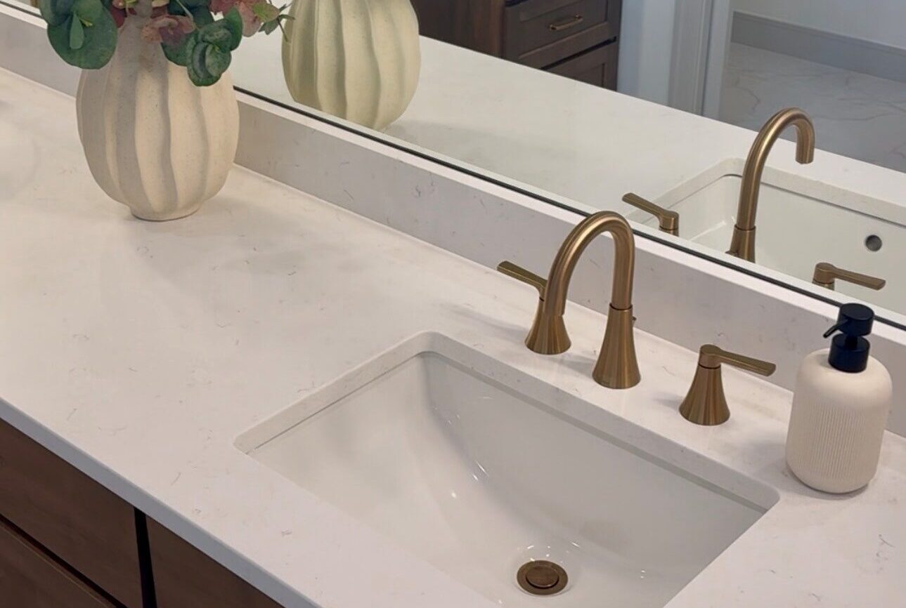 Modern bathroom vanity with gold faucet, white sink, two vases with greenery, and a soap dispenser.
