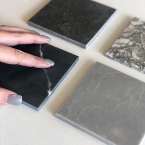 A hand with gray nails touches one of four square stone or tile samples on a light surface.