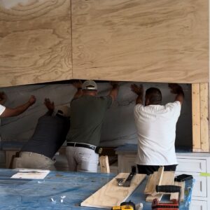FHBS install Bickimer Homes Three men install a large marble backsplash in a kitchen under unfinished wooden cabinets.